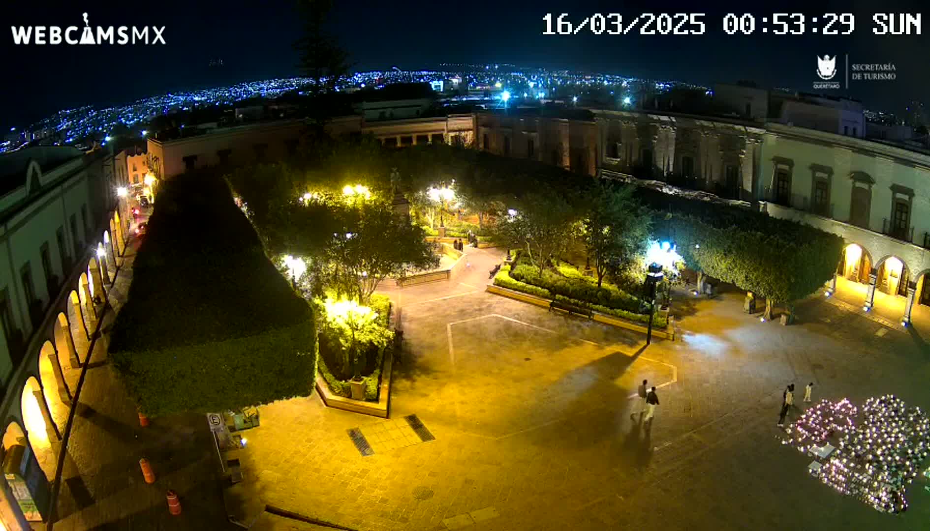 An illuminated plaza at night, surrounded by buildings, with a city skyline visible in the distance under a clear sky.