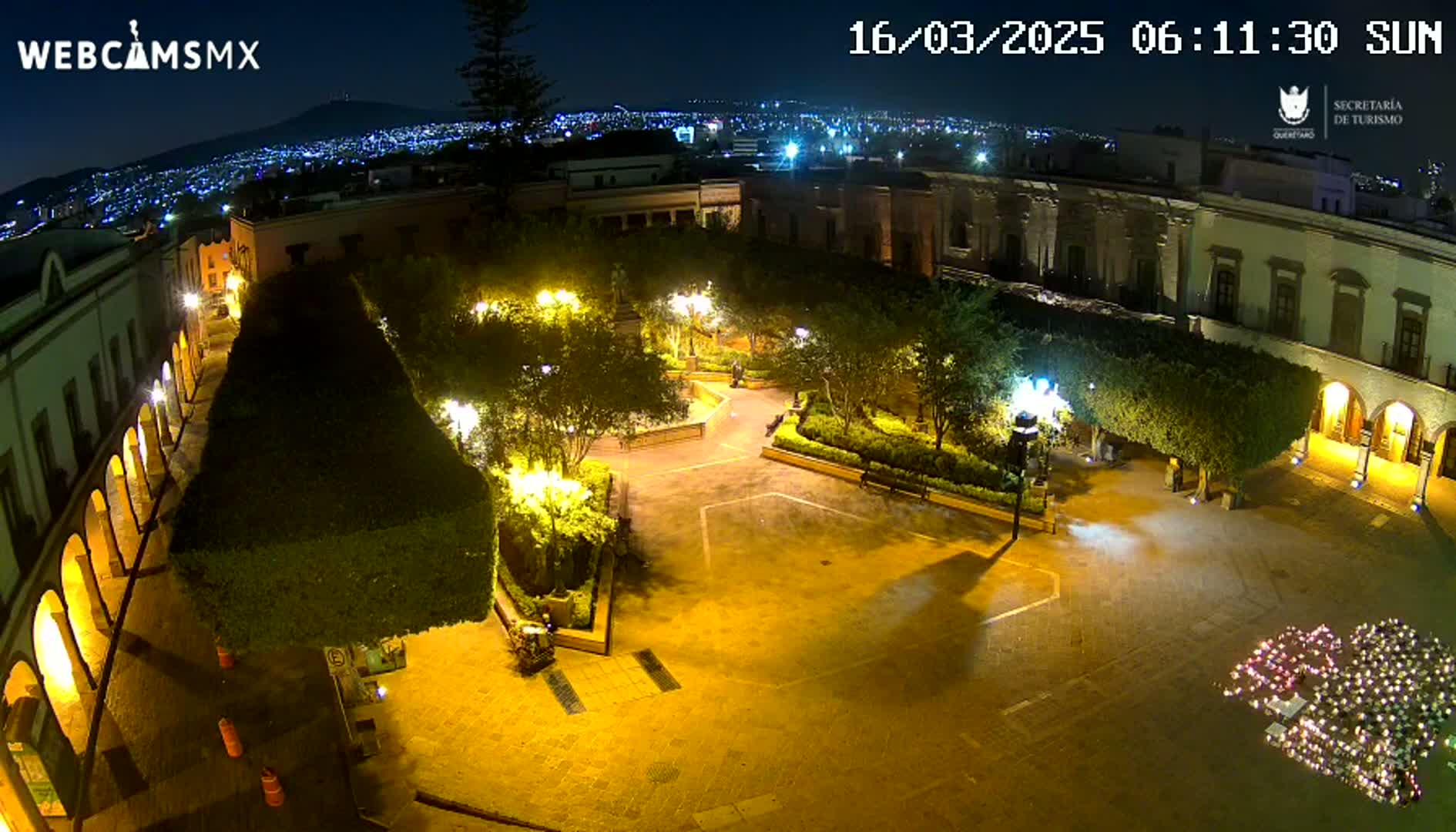 An illuminated town square at night, surrounded by buildings and lush greenery, with a distant city skyline visible under a clear sky.