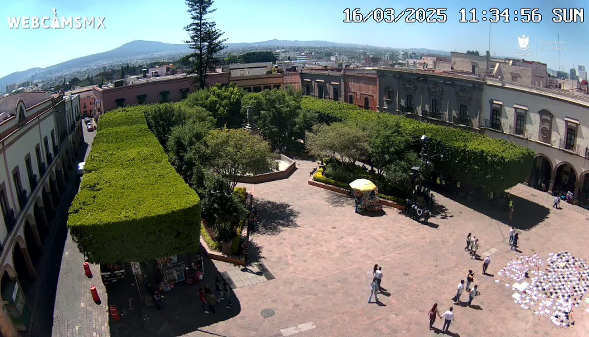 A sunny plaza with a central square surrounded by buildings and neatly trimmed hedges, numerous people walking around and a large collection of white papers laid on the ground.