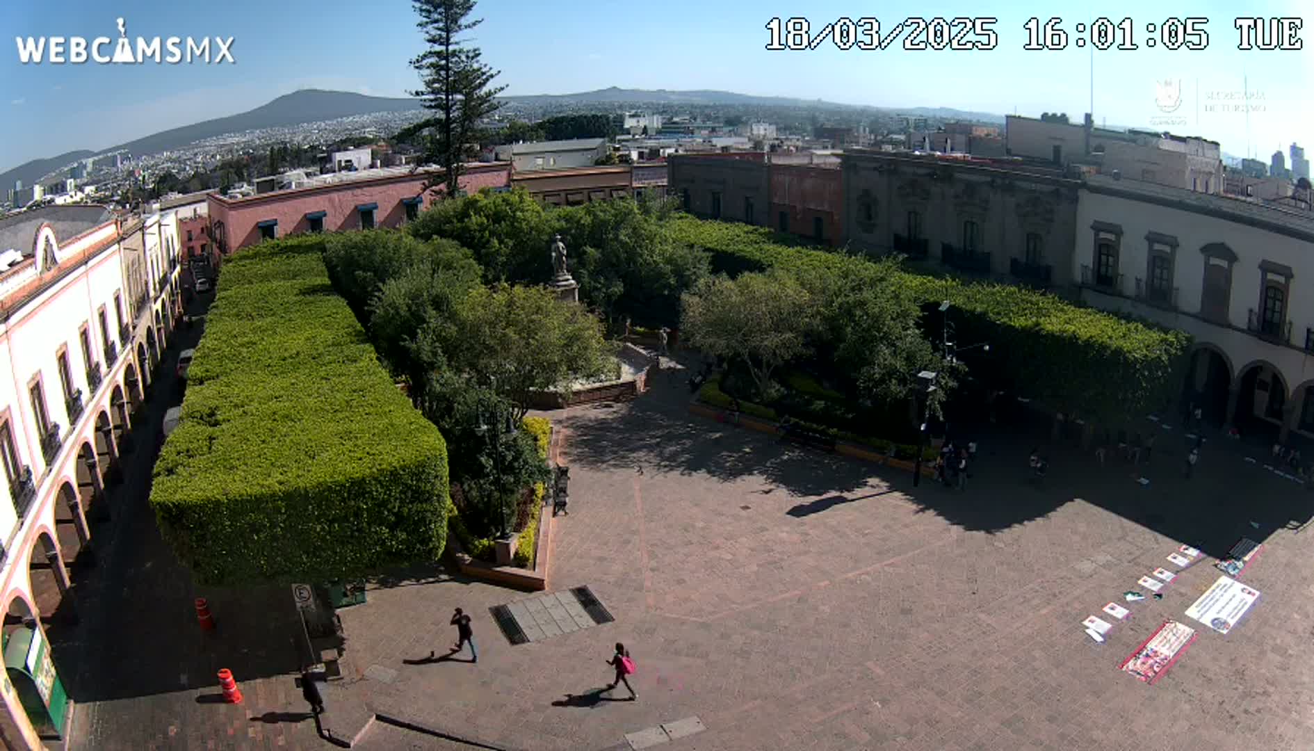 A high-angle view of a plaza with a central fountain and statue, surrounded by neatly trimmed hedges and colonial-style buildings, on a sunny day with a mountain visible in the distance.