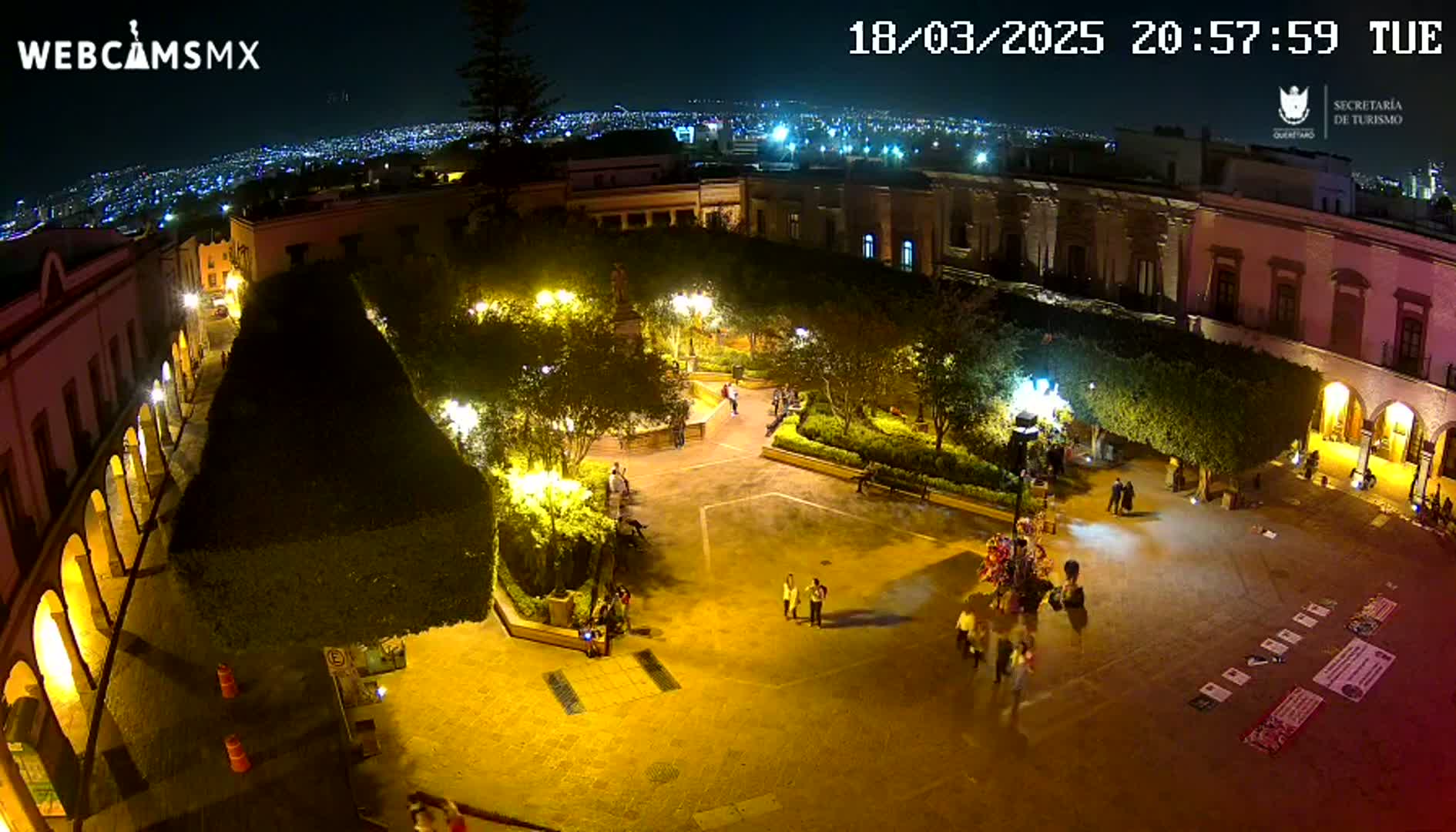An illuminated plaza at night, surrounded by buildings, with a few people walking around and a city skyline visible in the distance.