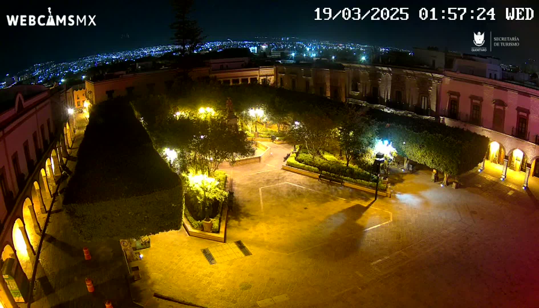 A nighttime aerial view of a town square, illuminated by streetlights, surrounded by buildings and lush, well-lit landscaping.