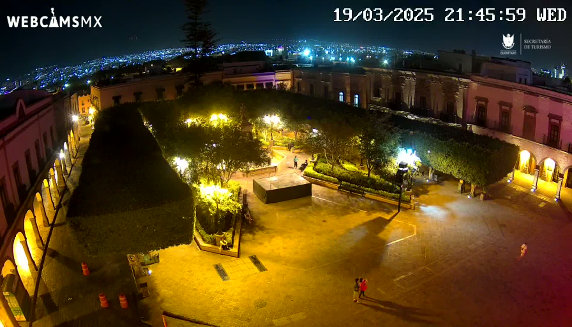 A nighttime aerial view of a brightly lit plaza surrounded by buildings, with a few people visible in the square and a city skyline visible in the distance under a clear sky.