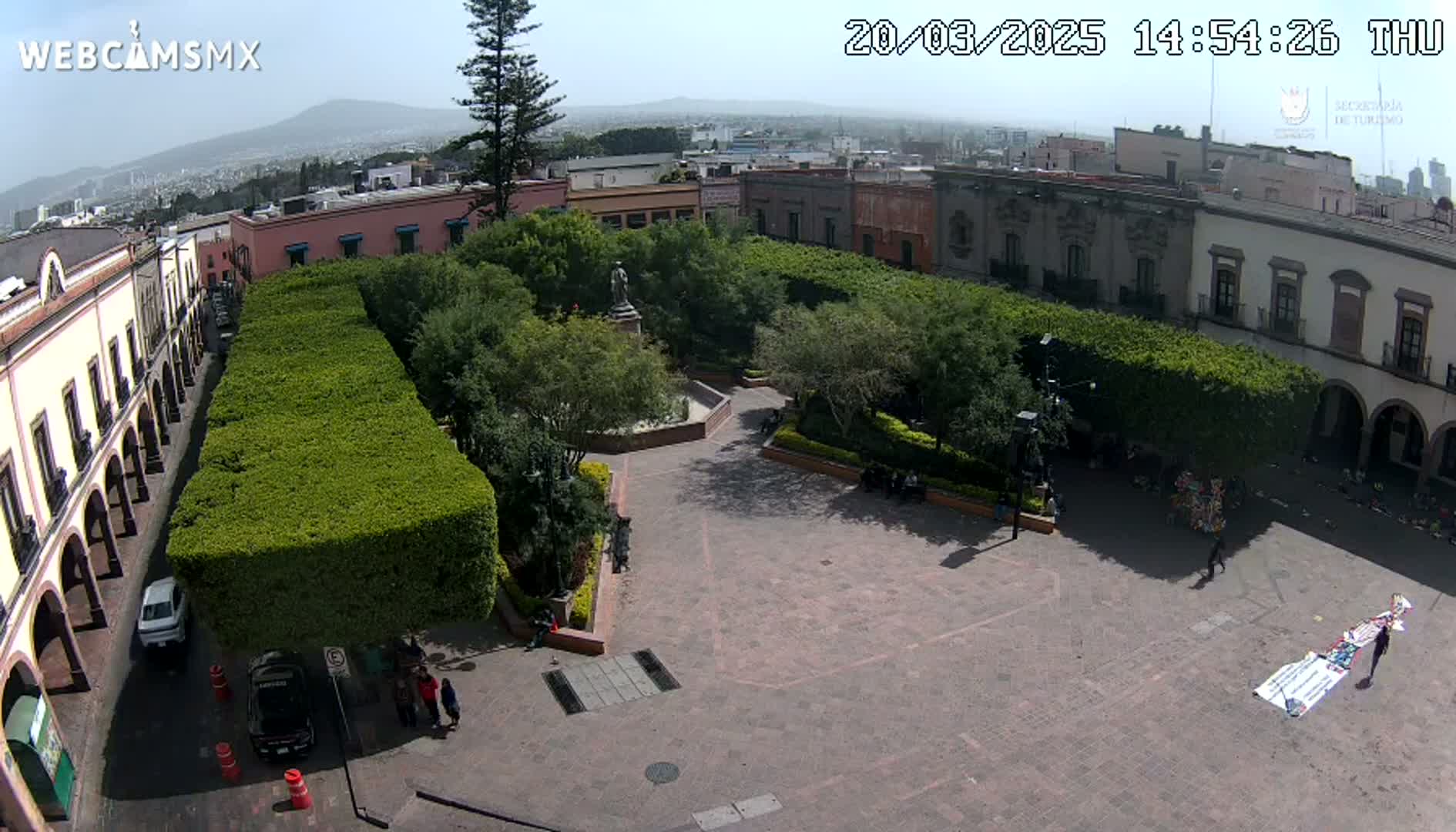 An aerial view of a town square with meticulously trimmed hedges surrounding a central plaza, under a clear, sunny sky.