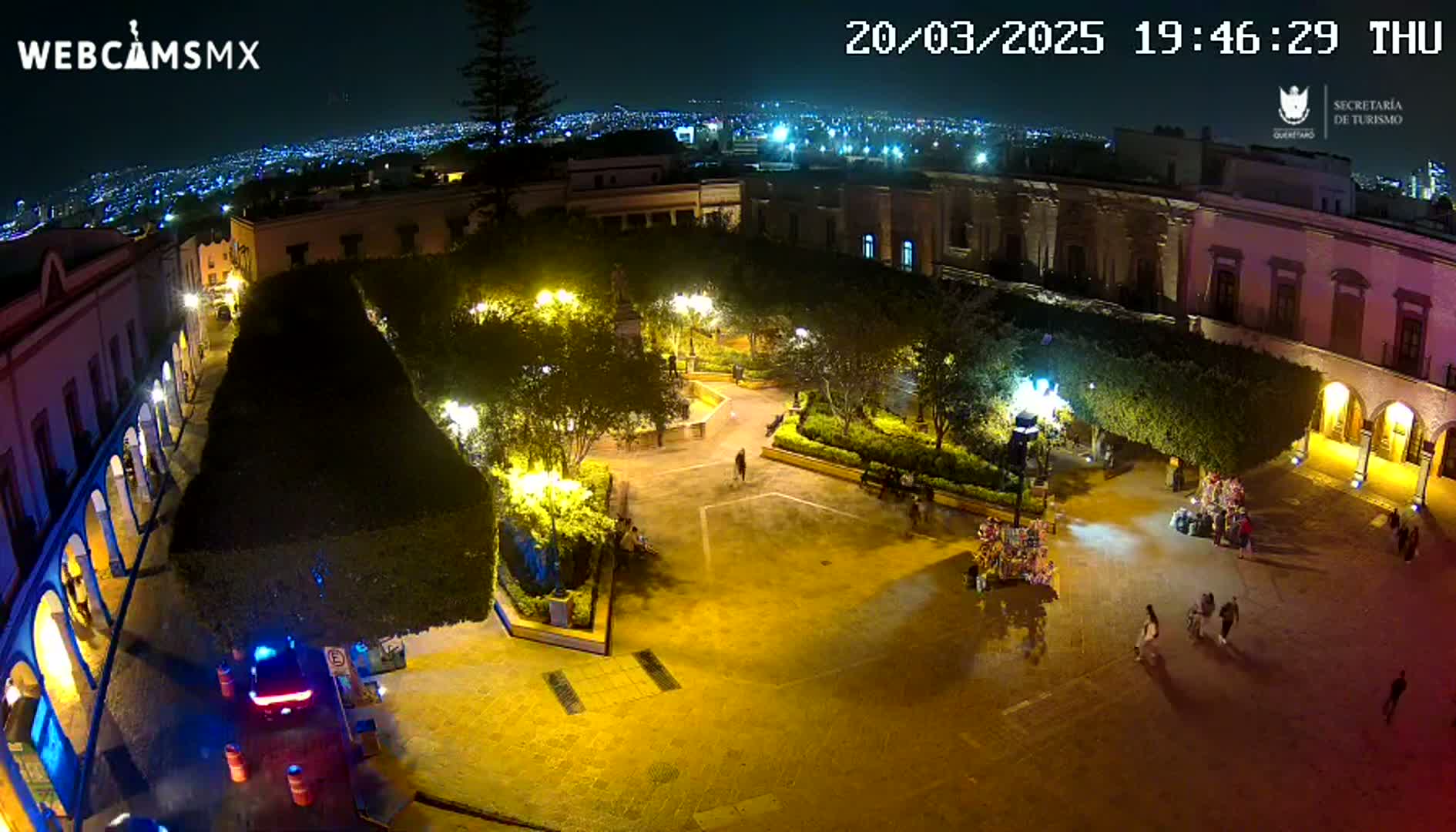 An illuminated plaza at night, surrounded by buildings, with a few people walking around and some vendors present.