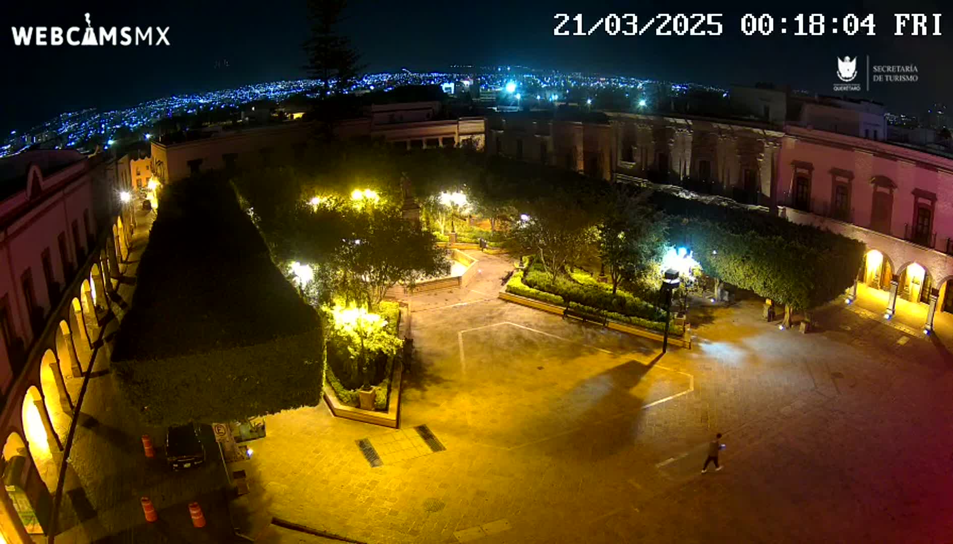 An illuminated plaza at night, surrounded by buildings, with a person walking across the square and a city visible in the distance.