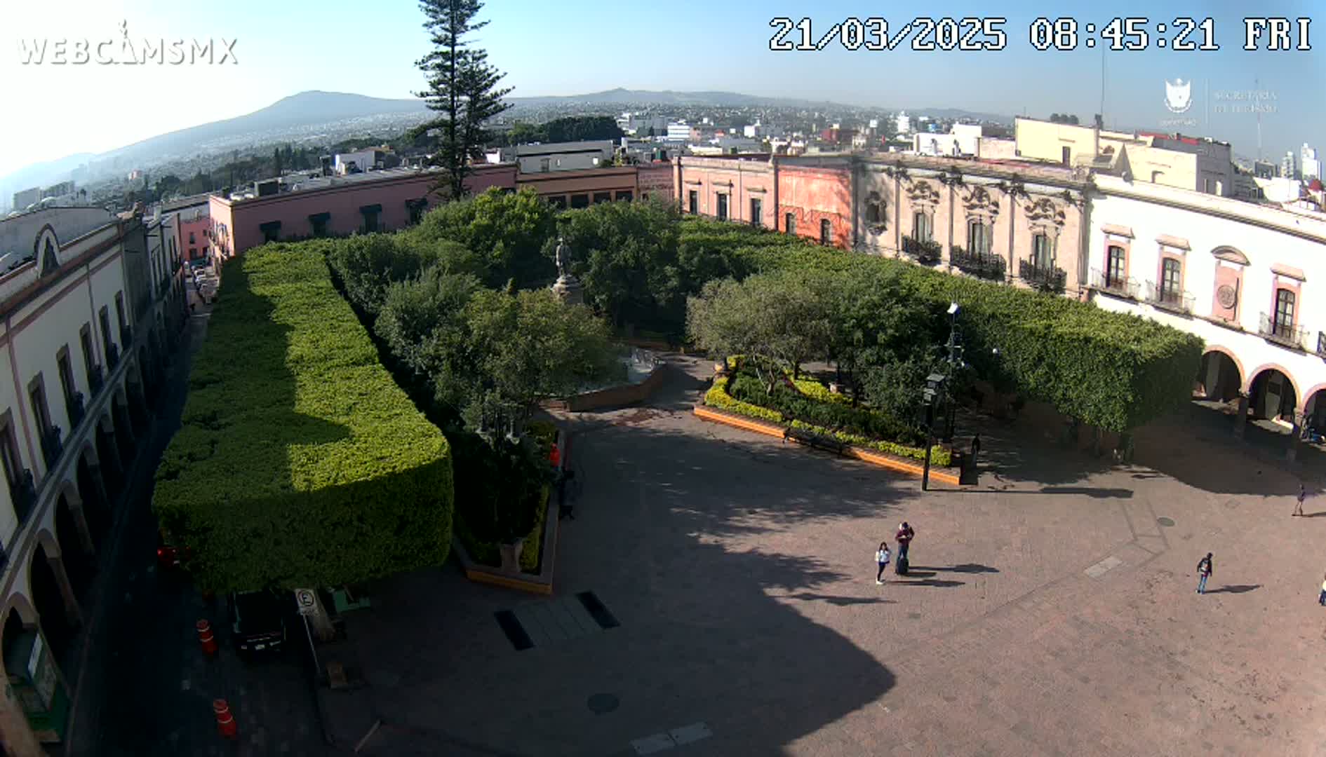 A mostly empty town square with manicured hedges, a fountain, and a statue, surrounded by buildings under a sunny sky.