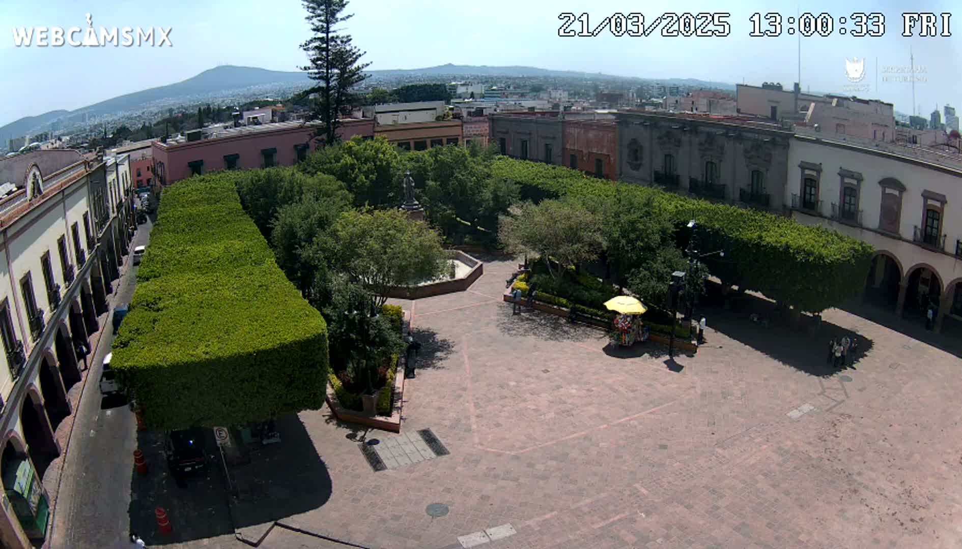 A sunny aerial view shows a town square with a central garden surrounded by trimmed hedges, buildings, and a few people.