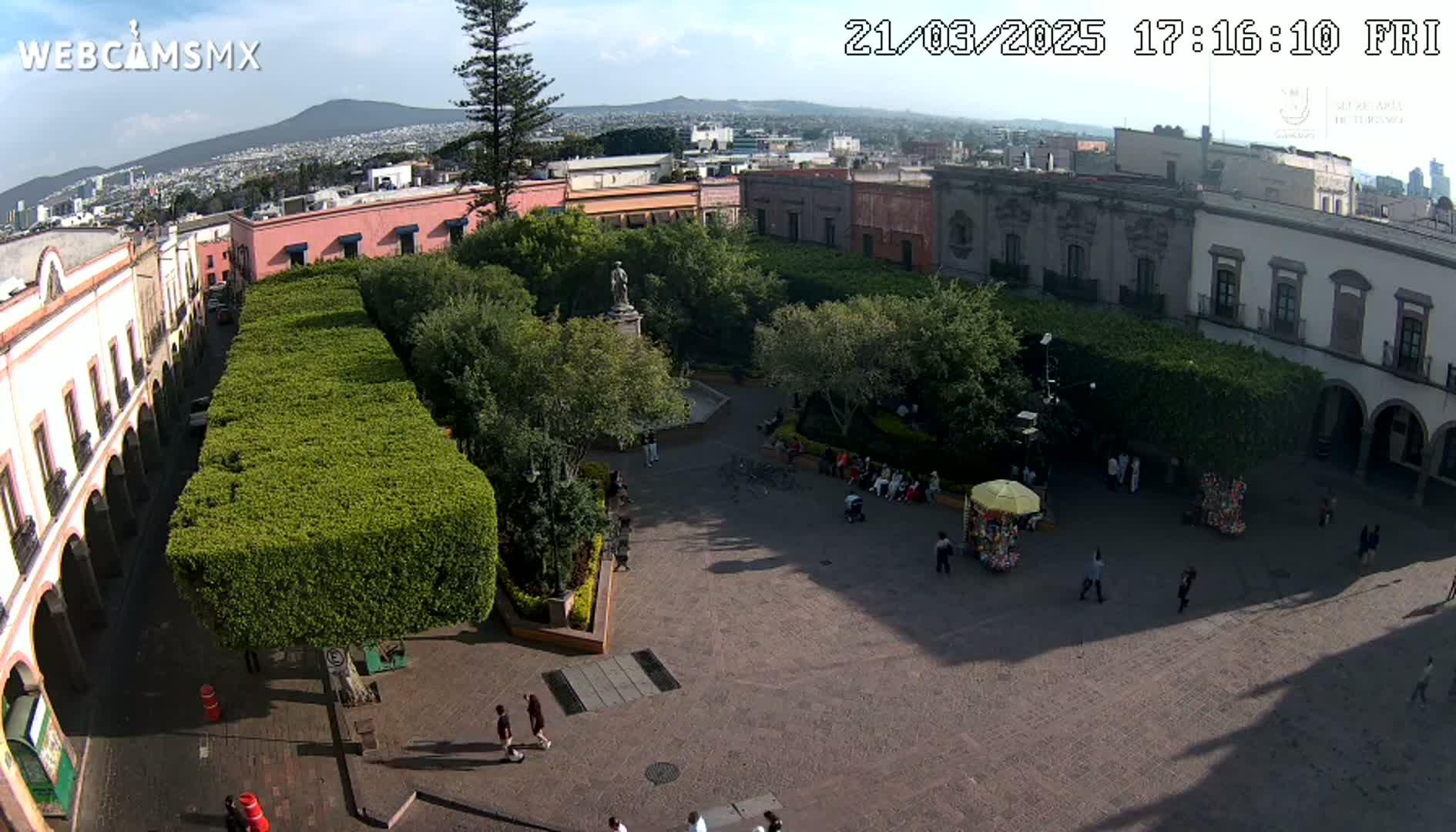 A high-angle view of a town square with green hedges, trees, a statue, and several people, under a mostly sunny sky.