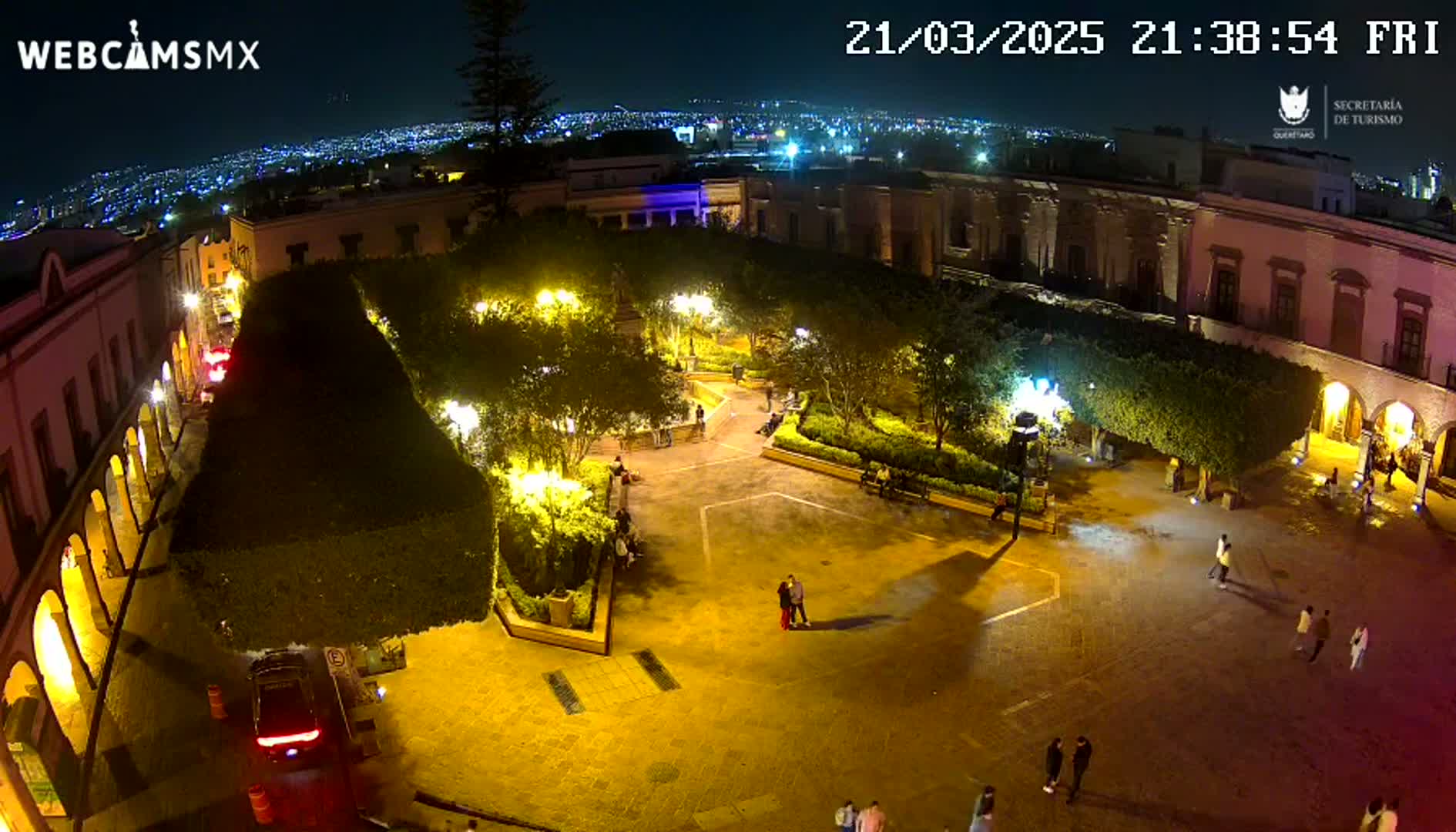 A nighttime aerial view of a town square, illuminated by streetlights, with several people walking around and cars parked nearby.