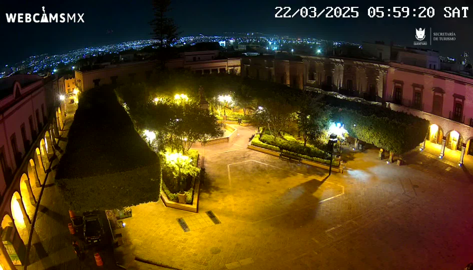 A nighttime aerial view of a plaza surrounded by buildings, illuminated by artificial lights, with a city skyline visible in the background under a clear sky.