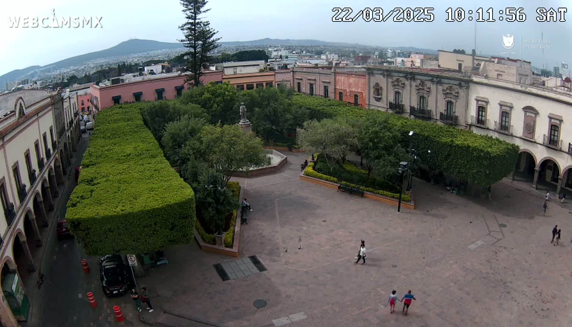 An aerial view of a town square with a central fountain, surrounded by buildings and neatly trimmed hedges, under an overcast sky.