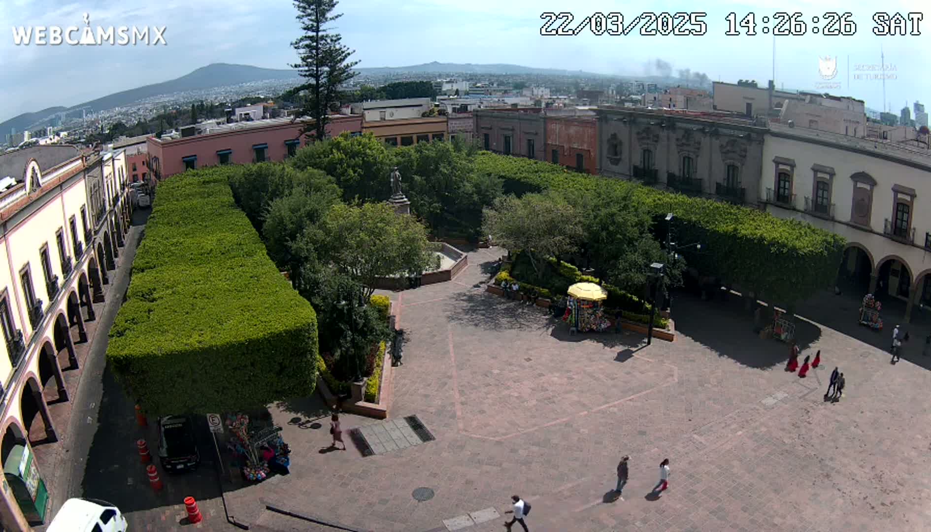 An aerial view shows a town square with neatly trimmed hedges, a central fountain, and several people walking around on a sunny day, with a distant mountain visible in the background.