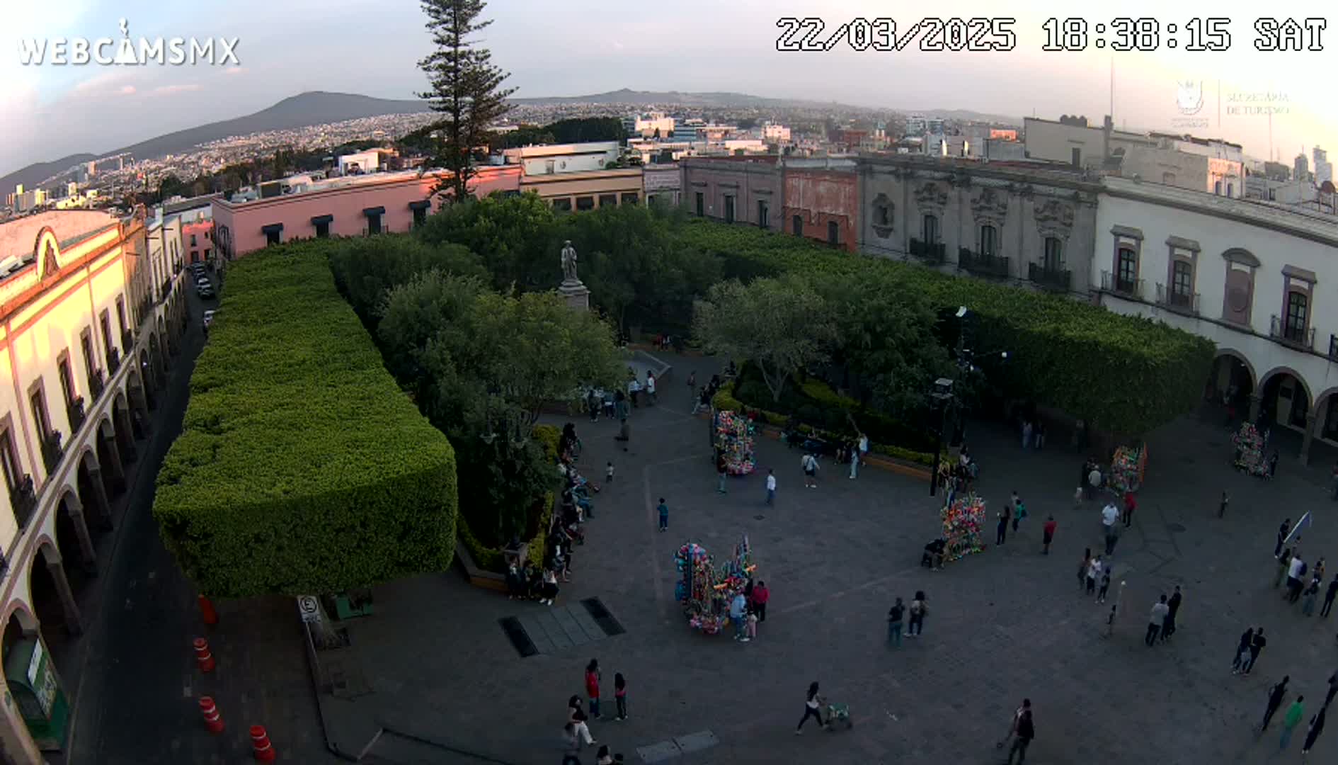 An aerial view shows a town square with people walking around and several vendors' stands, surrounded by buildings and lush green hedges under a partly cloudy evening sky.