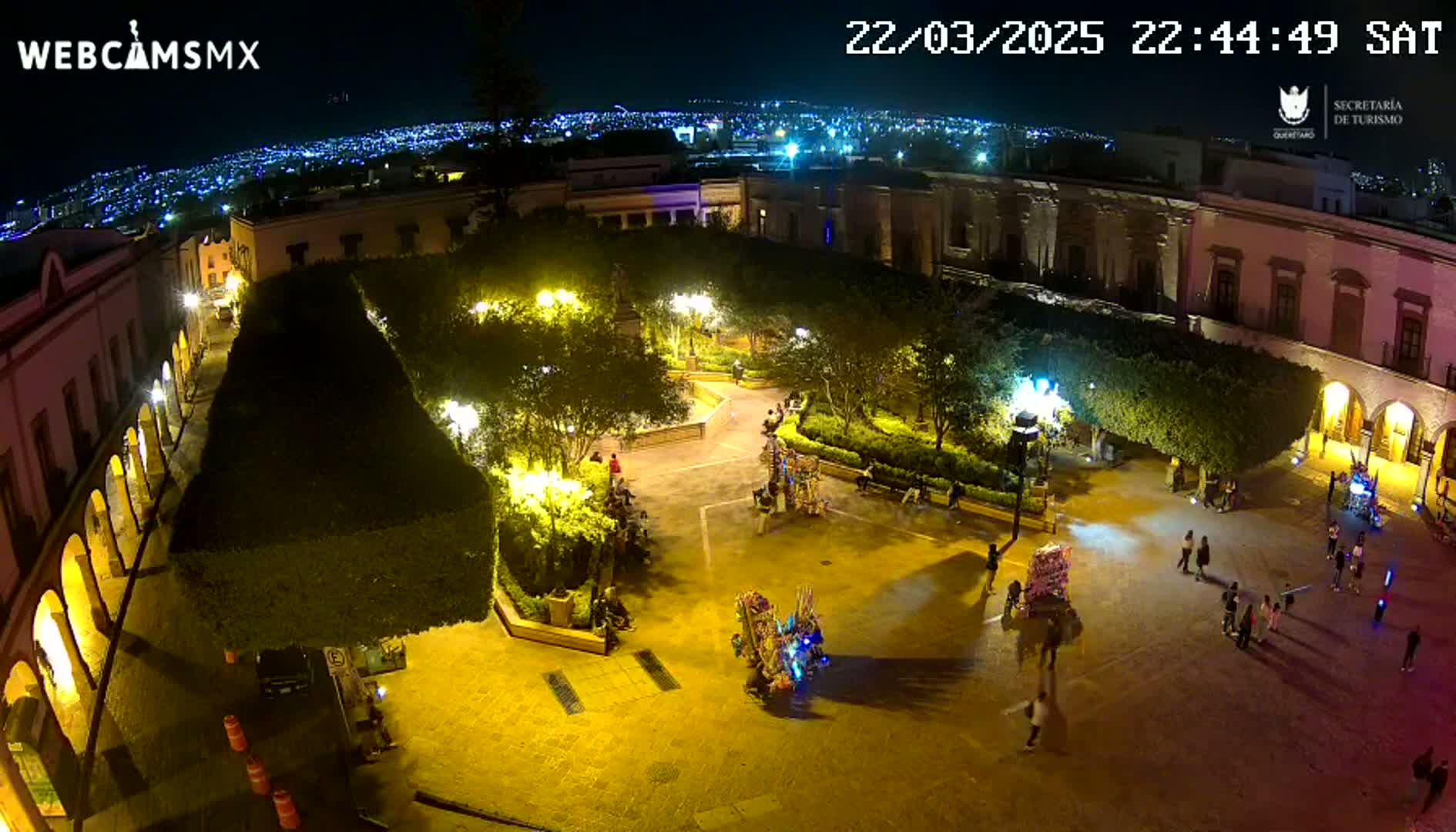 A nighttime, high-angle view of a town square, illuminated by streetlights, with people strolling around a plaza surrounded by buildings and lush greenery.