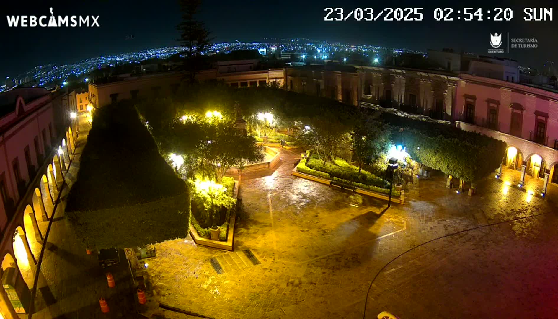 A nighttime, wet plaza is illuminated by streetlights, surrounded by buildings and lush greenery, with a city skyline visible in the distance.