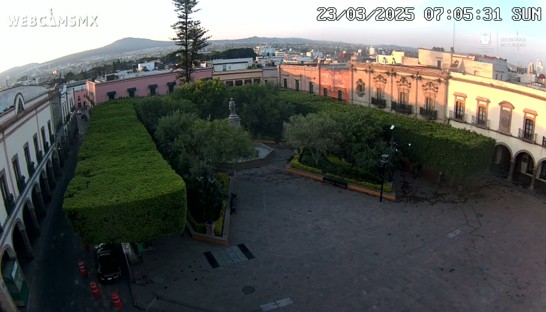 A paved plaza, surrounded by colonial-style buildings and neatly trimmed hedges, contains a central fountain and statue, under a clear, sunrise sky.