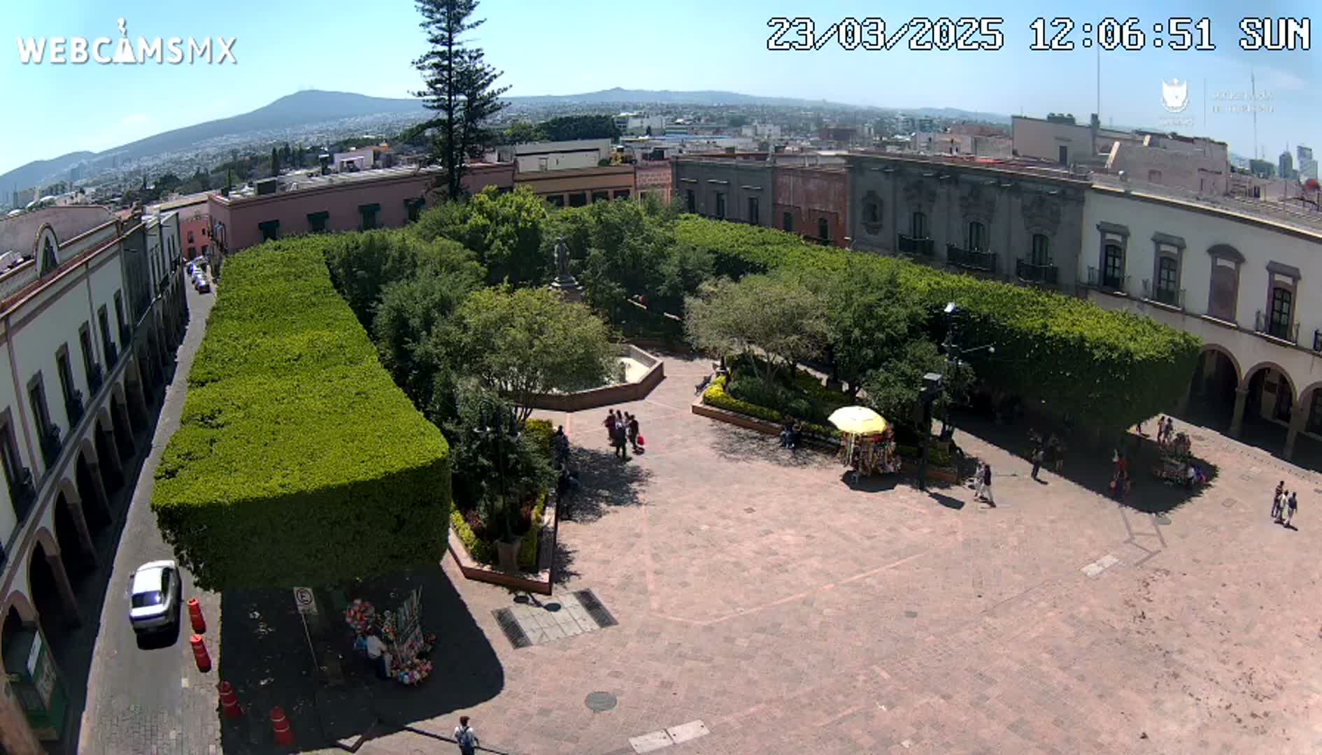 An aerial view of a town square on a sunny day, showing a paved plaza surrounded by buildings and lush green hedges, with a few people walking around.