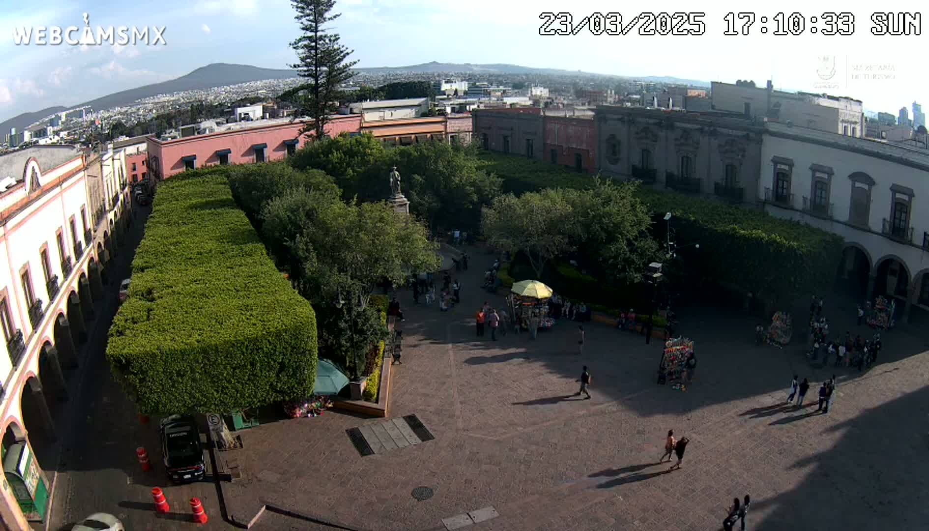 An elevated view of a town square, under sunny skies, shows a central plaza surrounded by buildings and lush greenery, with several people walking around.