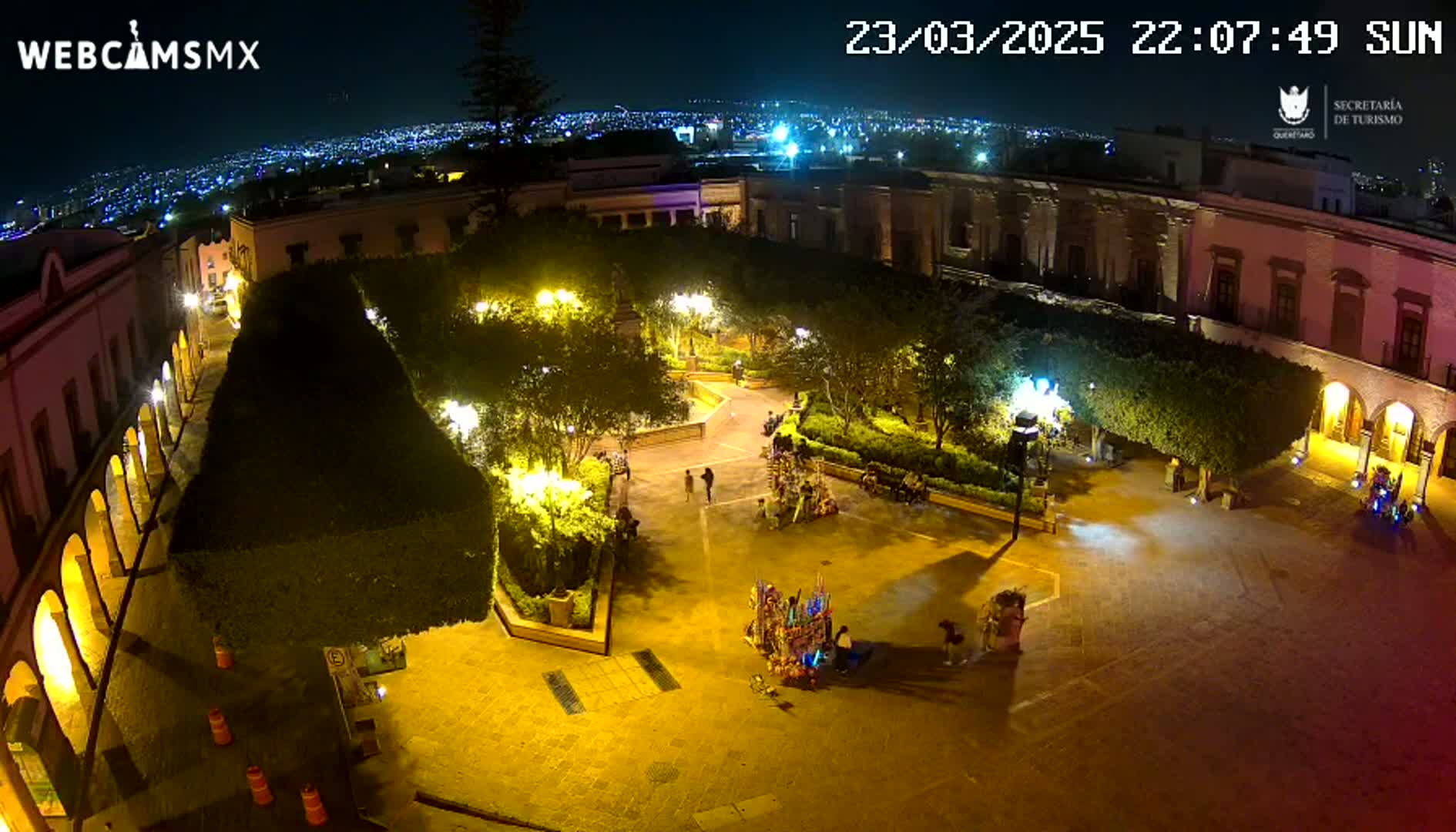 A nighttime aerial view of a town square, illuminated by artificial lights, with a few people present.