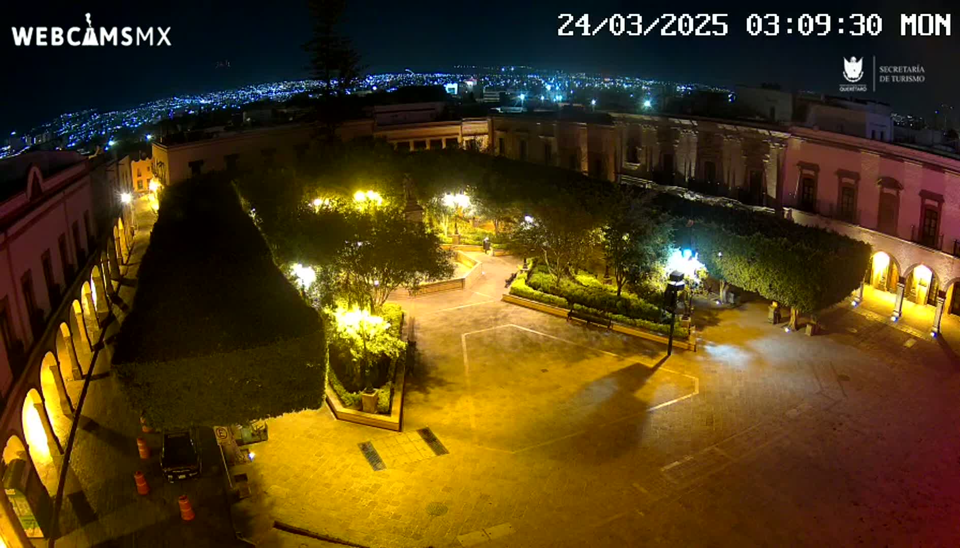 A nighttime view of a town square, illuminated by streetlights, surrounded by buildings and landscaping, under a clear sky.