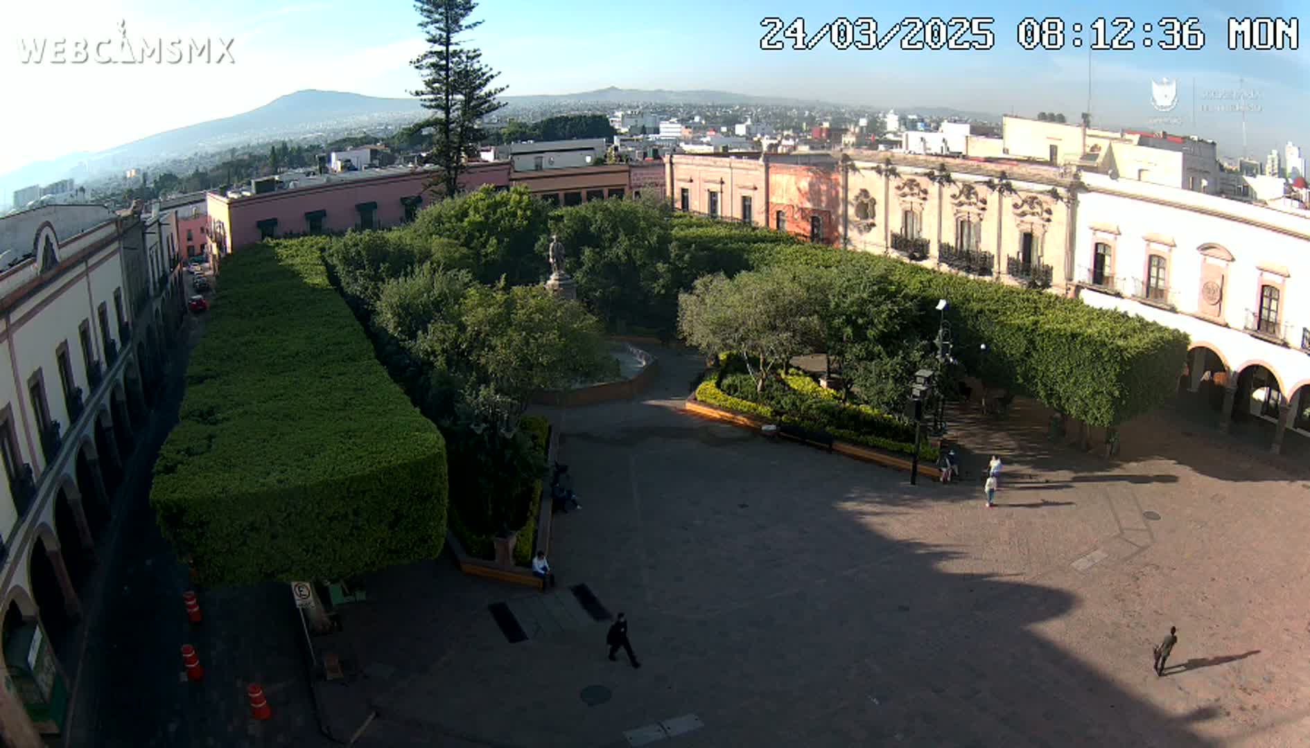 A sunny day reveals a town square surrounded by colonial-style buildings and neatly trimmed hedges, with a few people scattered throughout.