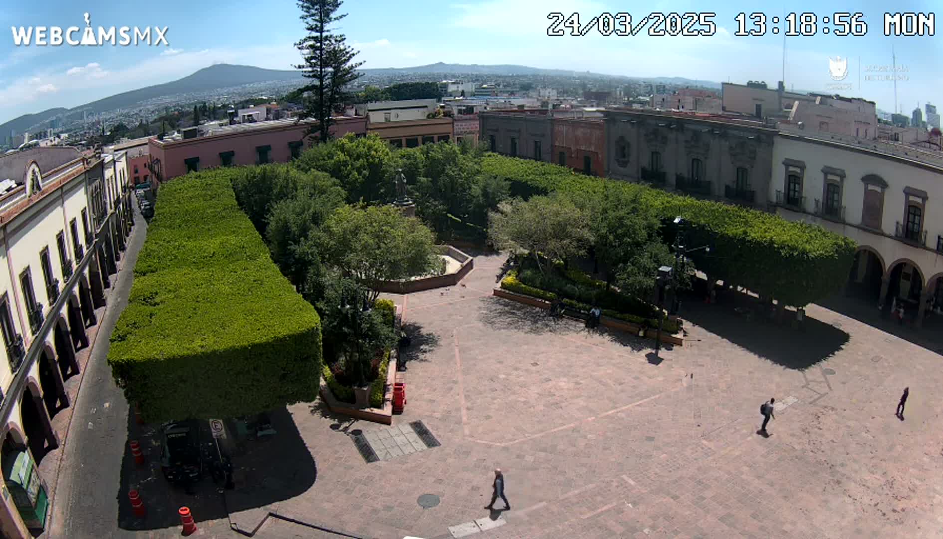 An aerial view of a town square with a central garden surrounded by buildings and trimmed hedges, on a sunny day.