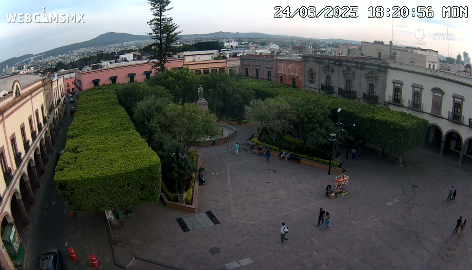 An aerial view of a town square with a statue, fountain, and several people, surrounded by buildings and lush green hedges under a mostly clear sky.