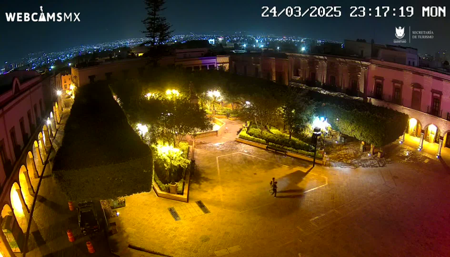 A nighttime aerial view of a plaza, illuminated by streetlights, surrounded by buildings and landscaped gardens, under a clear sky.