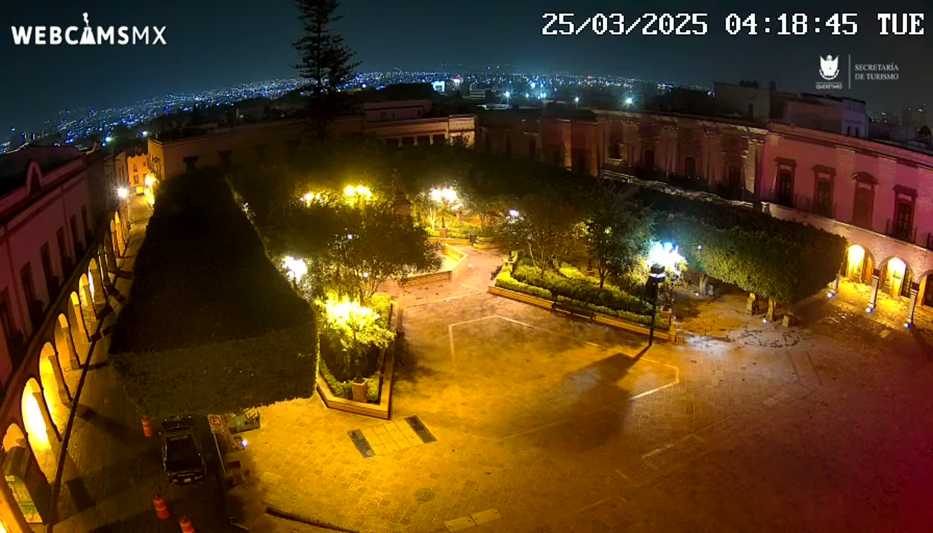 An illuminated plaza at night, surrounded by buildings, with landscaped areas and a few vehicles parked nearby.