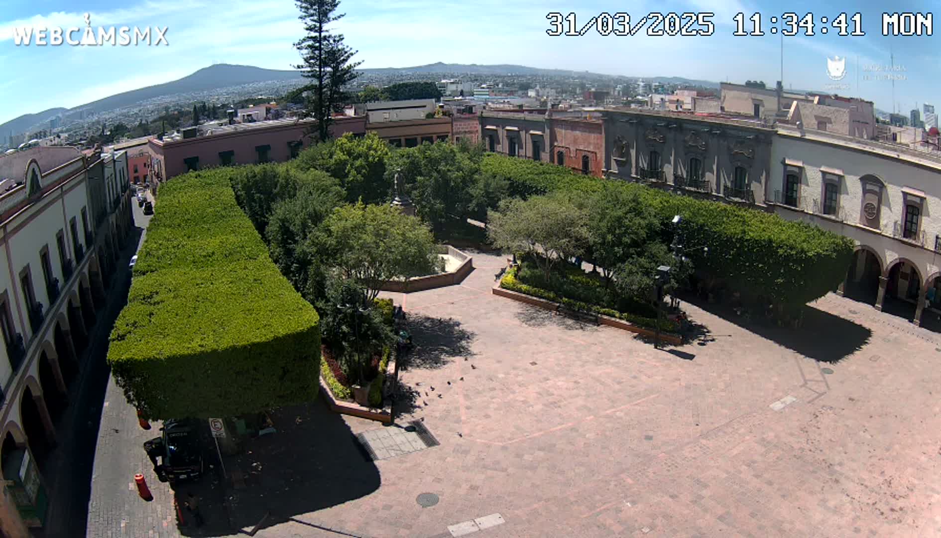 An elevated view of a plaza with a central paved area surrounded by trimmed hedges and buildings under a sunny sky, showing a distant mountain range.