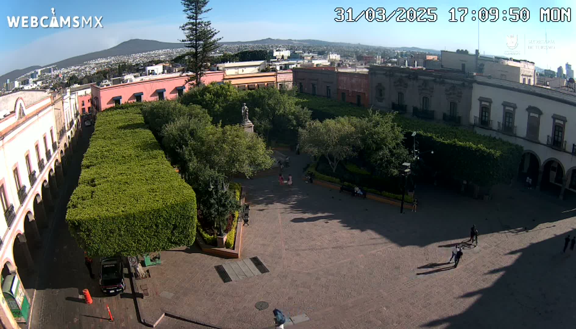 An aerial view of a town square with a central garden, surrounded by colonial-style buildings under a clear, sunny sky.