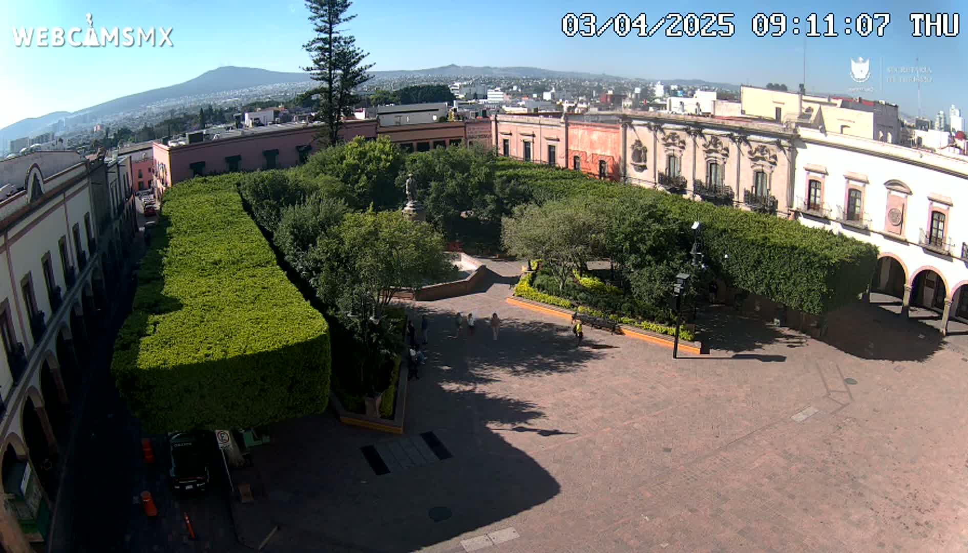 An aerial view of a town square with a central plaza surrounded by buildings and hedges, on a sunny day.