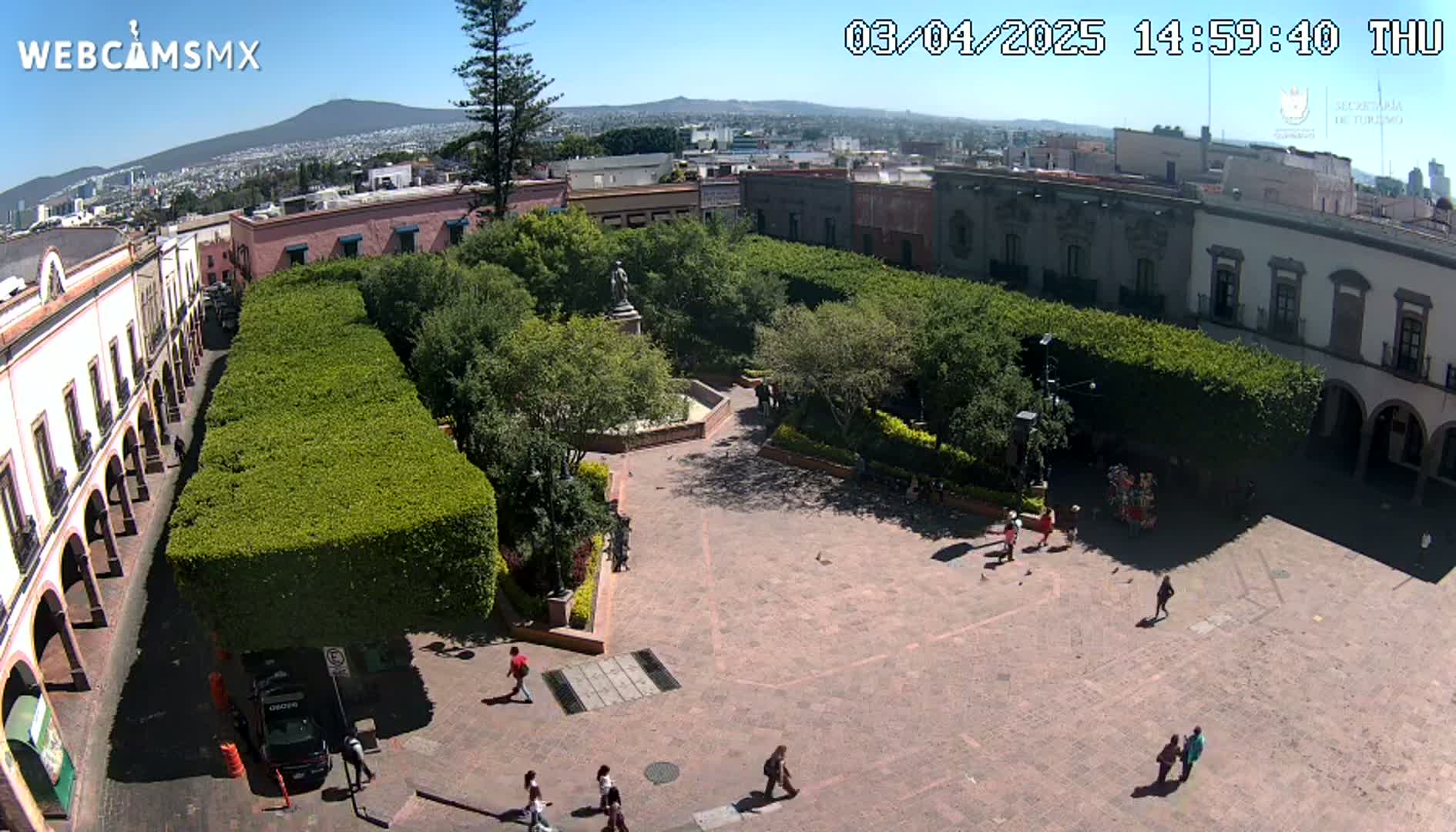 An aerial view of a plaza with a central garden featuring trimmed hedges and a statue, surrounded by buildings under a sunny sky.