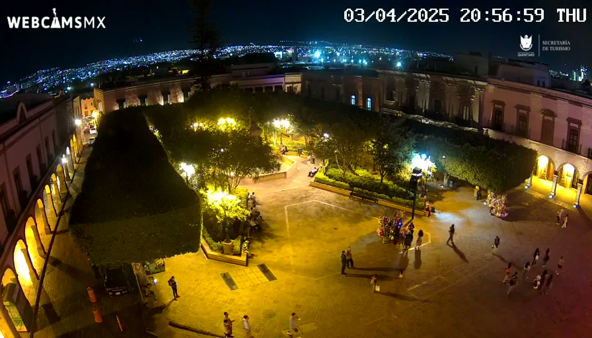 A nighttime aerial view of a town square, illuminated by streetlights, with people walking around and a city skyline visible in the distance.