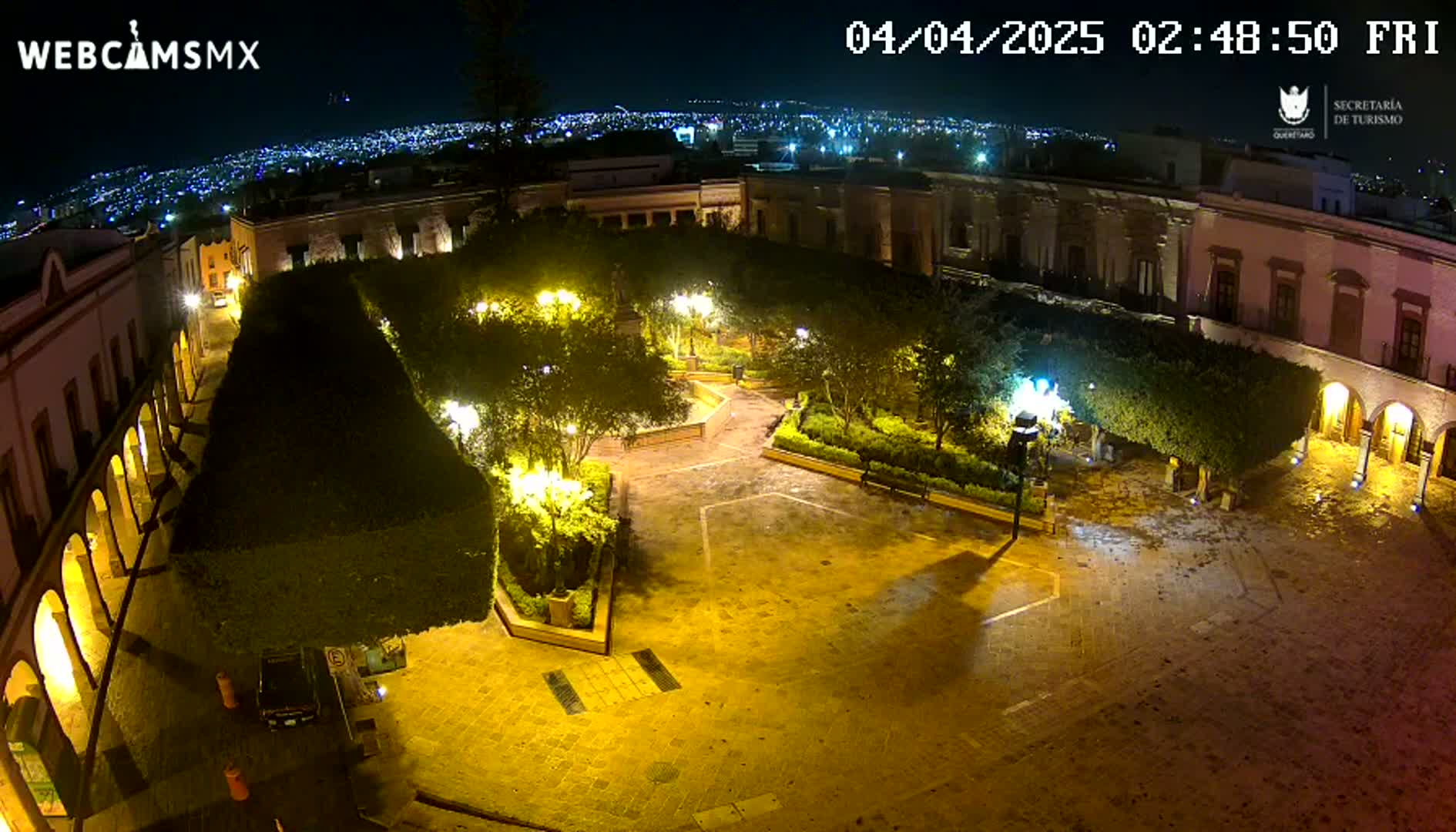 A nighttime, high-angle view of a plaza surrounded by buildings, illuminated by streetlights, with a city skyline visible in the distance.