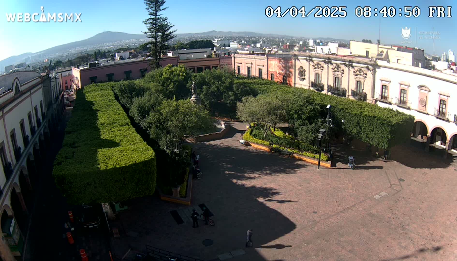 A paved plaza surrounded by colonial-style buildings and lush green hedges, under a clear sunny sky, is viewed from above.