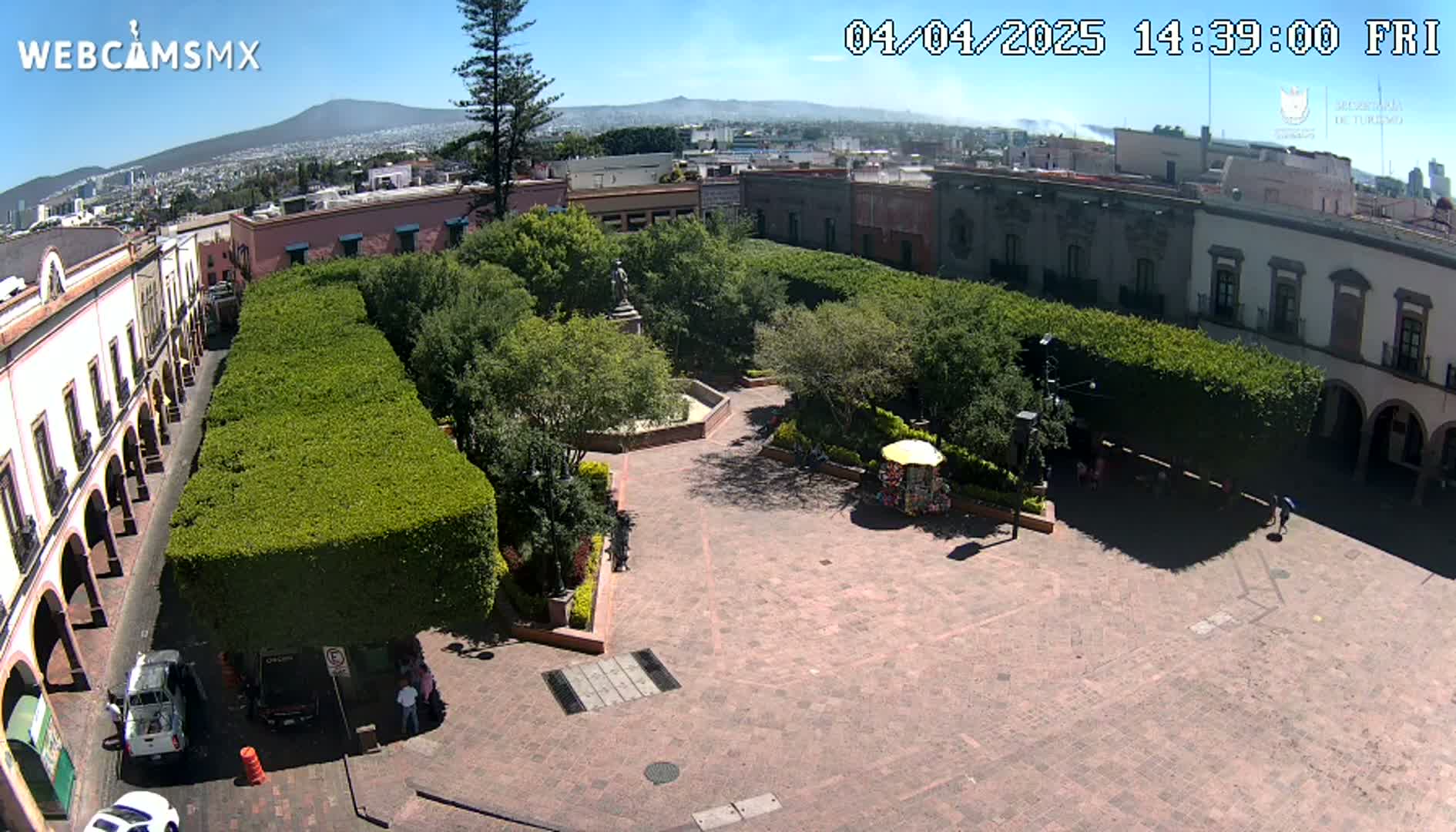 A paved plaza with a central garden surrounded by trimmed hedges, colonial-style buildings, and a distant mountain range under sunny skies.