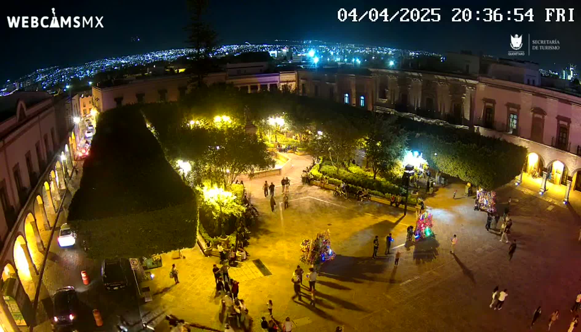 A brightly lit town square at night, surrounded by buildings, with people gathered and city lights visible in the distance under a clear sky.