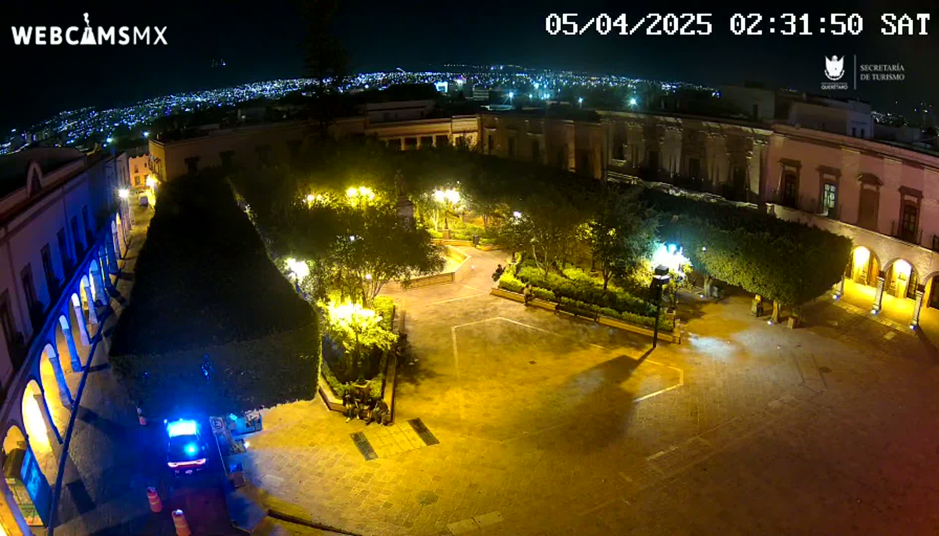 A nighttime aerial view of a town square, illuminated by streetlights, with a police vehicle parked near the edge of the square and a city skyline visible in the distance.