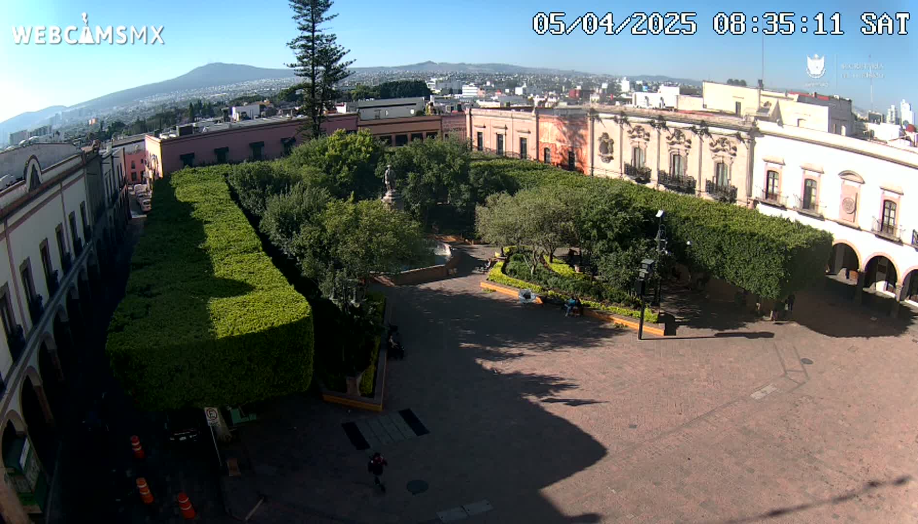 A sunny, high-angle view of a town square with a central park surrounded by low buildings,  tall hedges, and a few people.