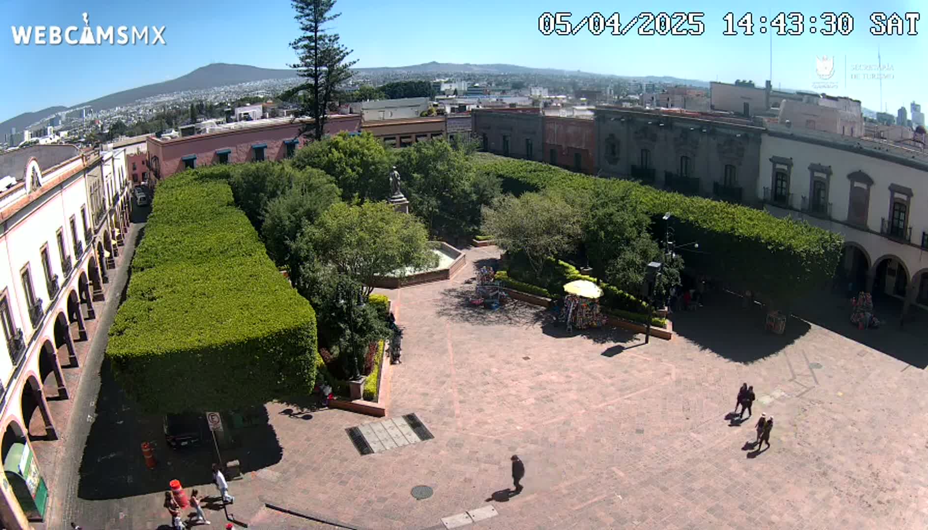 A sunny aerial view shows a plaza with a central fountain, surrounded by trimmed hedges, buildings, and a few people walking.