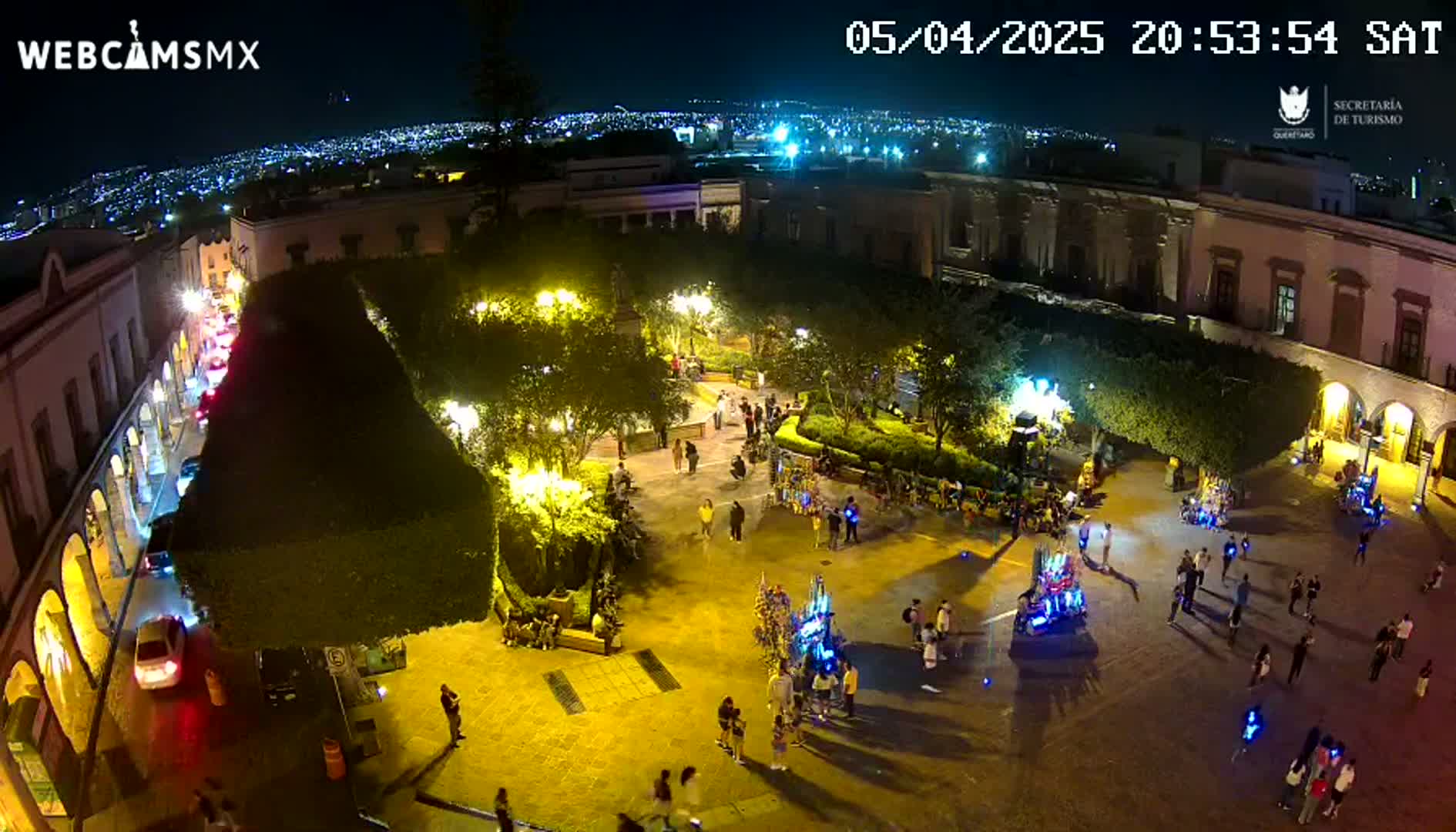 A brightly lit town square at night, filled with people and surrounded by buildings, overlooks a city skyline under a clear sky.
