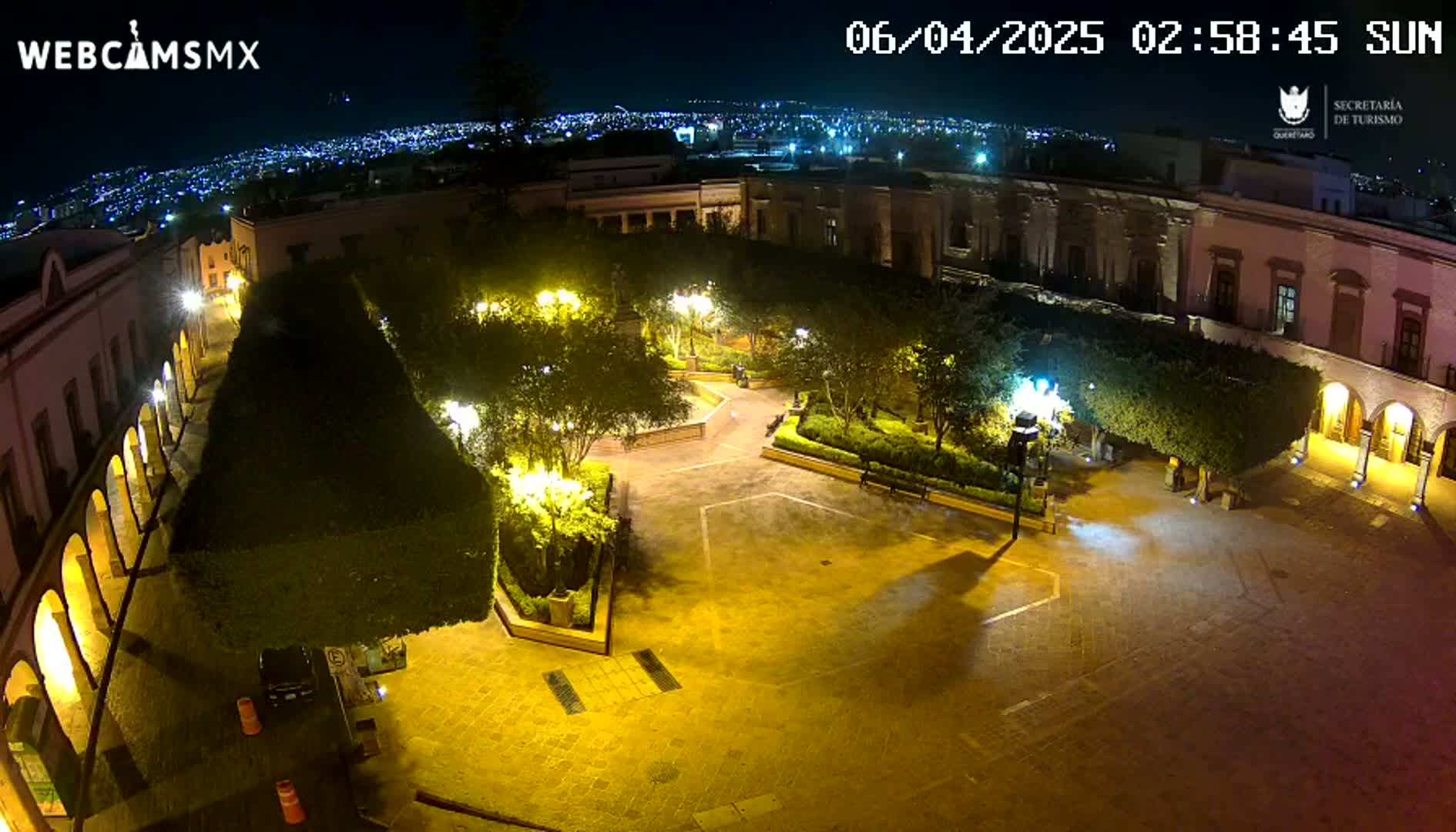 A nighttime aerial view of a town square, illuminated by streetlights, with buildings surrounding a central plaza containing neatly trimmed shrubs and trees.
