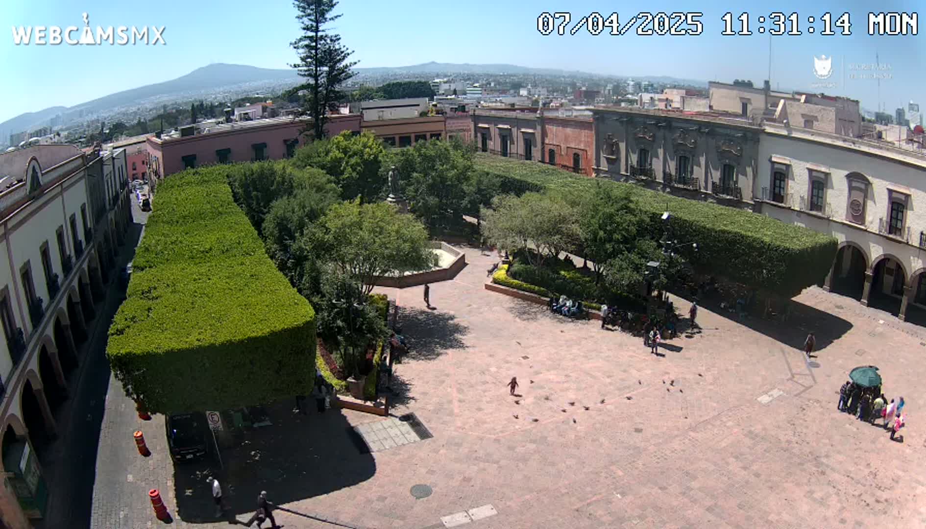 A sunny day reveals a town square with a central fountain, surrounded by trimmed hedges, buildings, and a few people strolling.