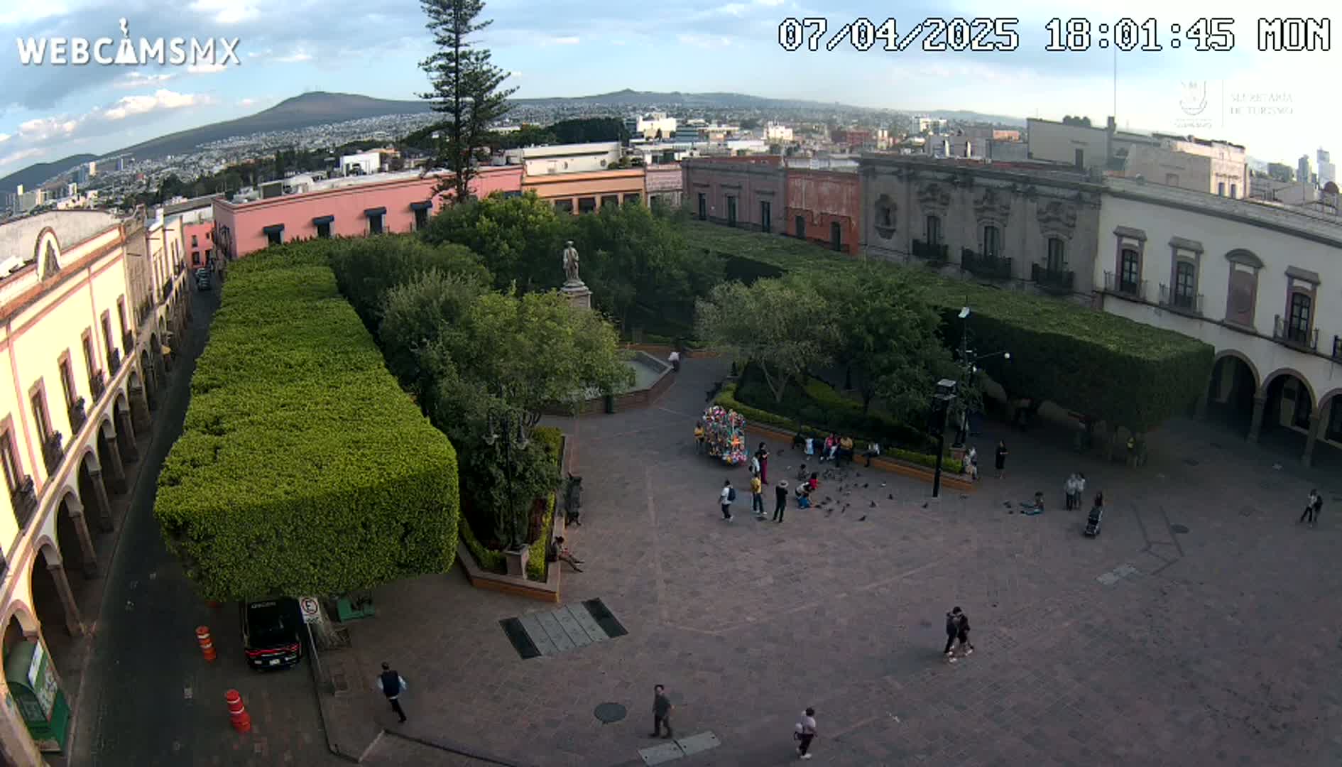A high-angle view shows a town square with a statue in the center, surrounded by buildings and trimmed hedges under a partly cloudy sky.