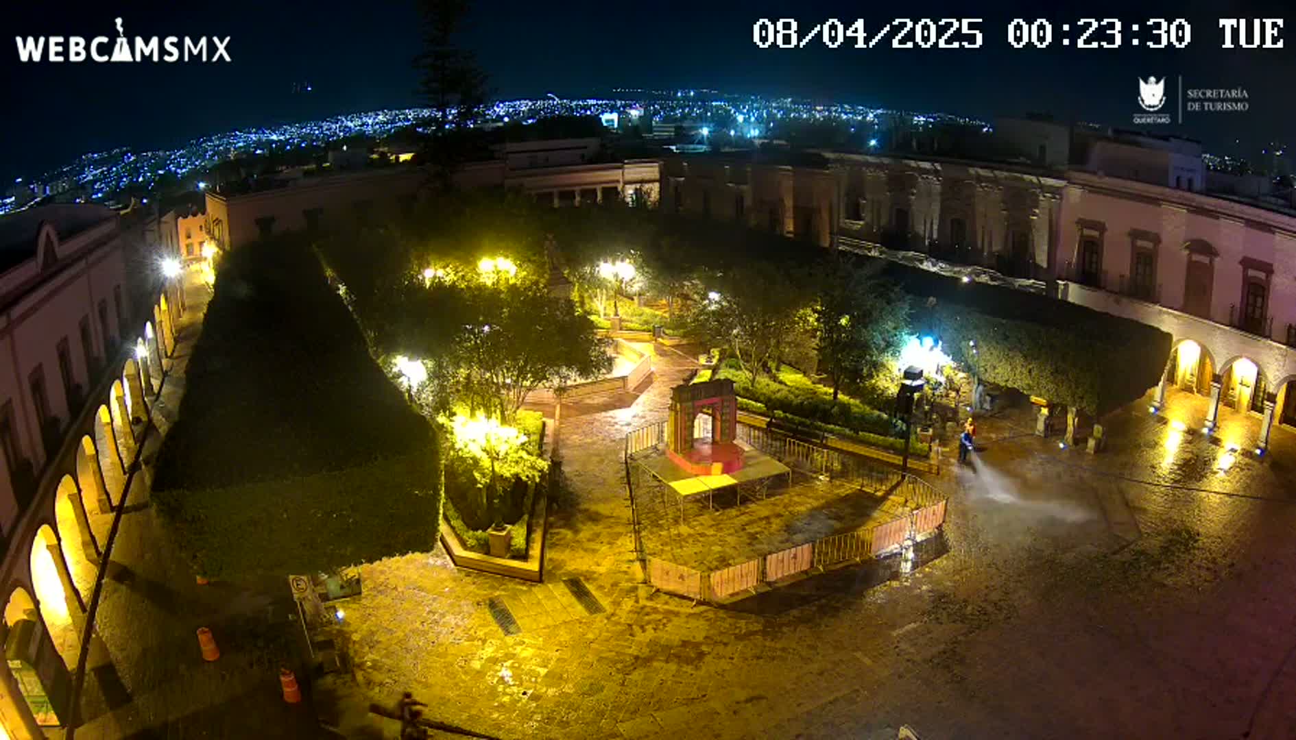 A nighttime overhead view of a plaza, illuminated by streetlights, with a person cleaning the wet pavement.