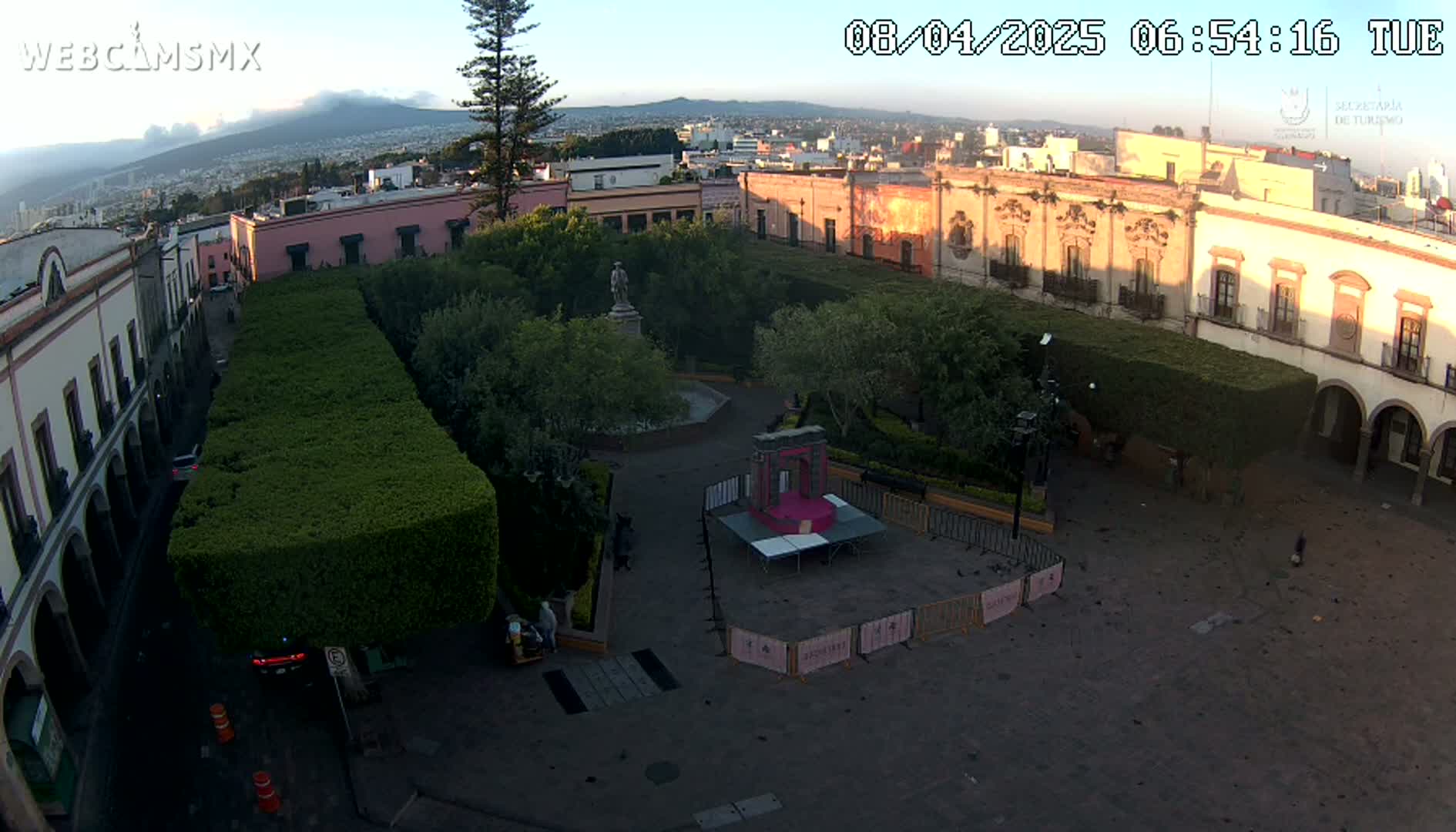 An aerial view of a plaza in a city at sunrise, showing a central square with a statue, neatly trimmed hedges, and colonial-style buildings surrounding it, with a distant mountain range visible under a clear sky.