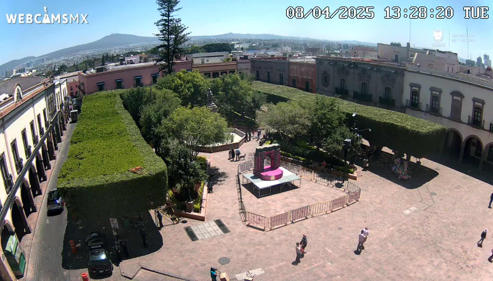A sunny aerial view of a town square with a stage set up, surrounded by buildings and trimmed hedges, under a clear sky.