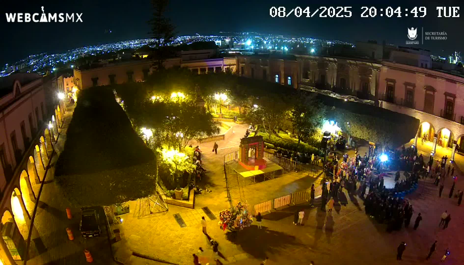 A nighttime aerial view shows a town square with people gathered around a central stage, illuminated by streetlights against a backdrop of a city skyline.